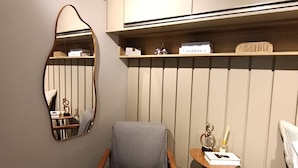 A compact room with a grey chair, a small table with a lamp, a mirror, and a bed with a white cover.