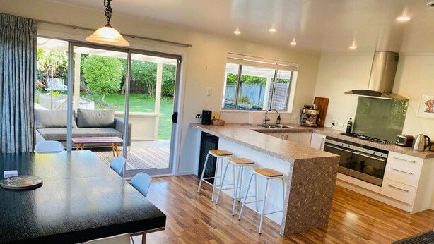 A modern kitchen with a central island, bar stools, a dining area with a table and chairs, and a sliding glass door leading to a patio. - Spacious 4 BRM home with spa and large garden (Taupō)