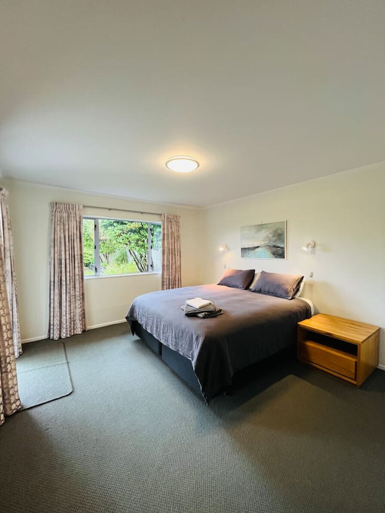 A bedroom with a bed, a bedside table, a window with curtains, and a painting on the wall.