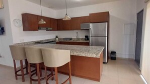 A kitchen with a granite countertop, wooden bar stools, a stainless steel refrigerator, and a wall-mounted clock. - Cozy 2-bedroom apartment in Cabo San Lucas. (Cabo San Lucas)