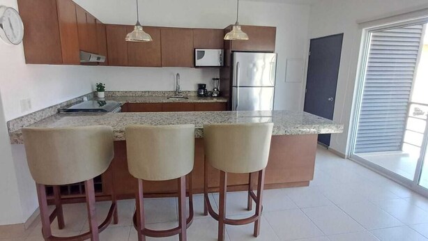 A kitchen with a granite countertop, stainless steel appliances, wooden cabinets, and three bar stools. - Cozy 2-bedroom apartment in Cabo San Lucas. (Cabo San Lucas)