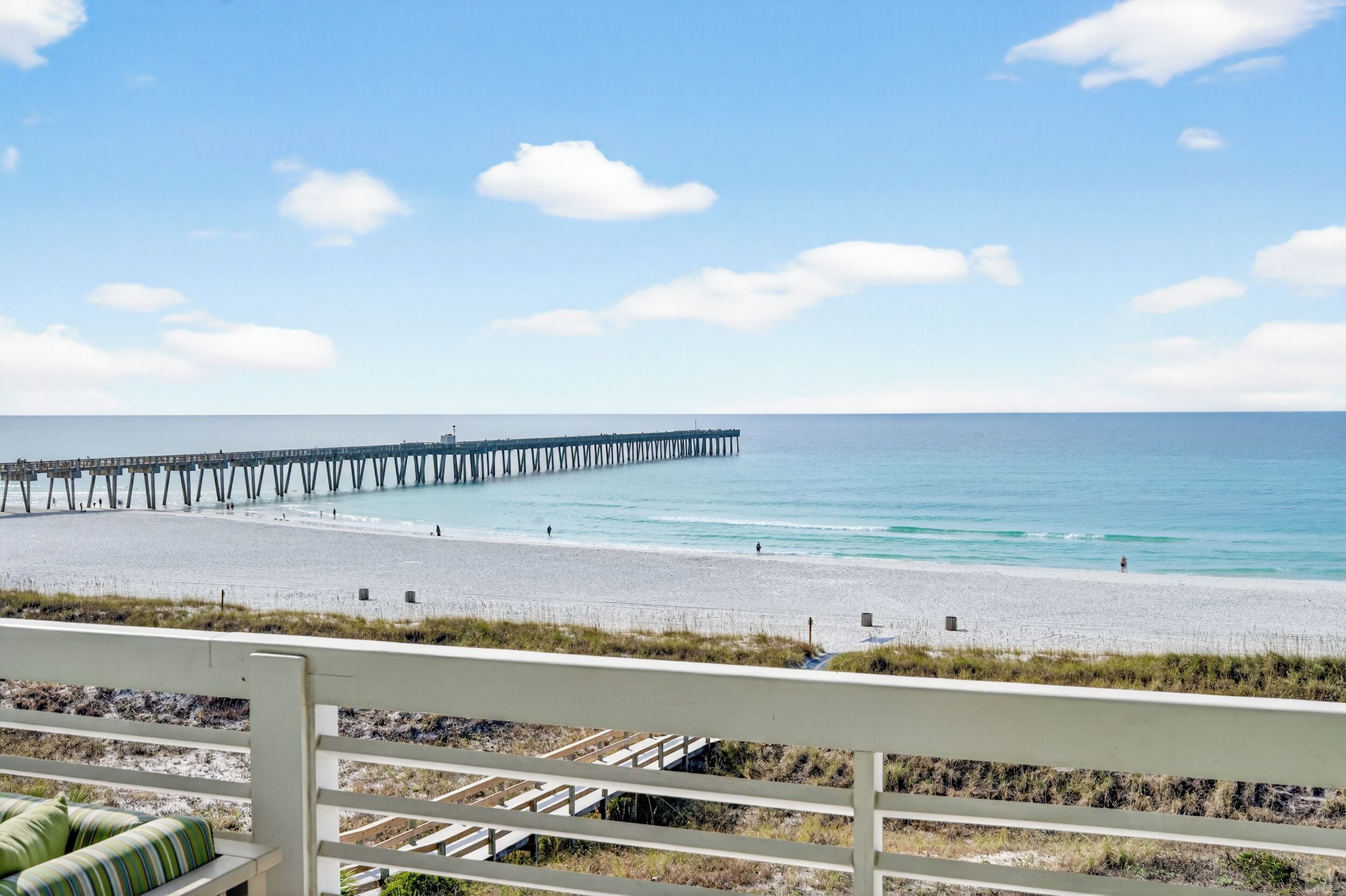 A beach with a pier extending into the water, a sandy shore, and a grassy area with a bench.