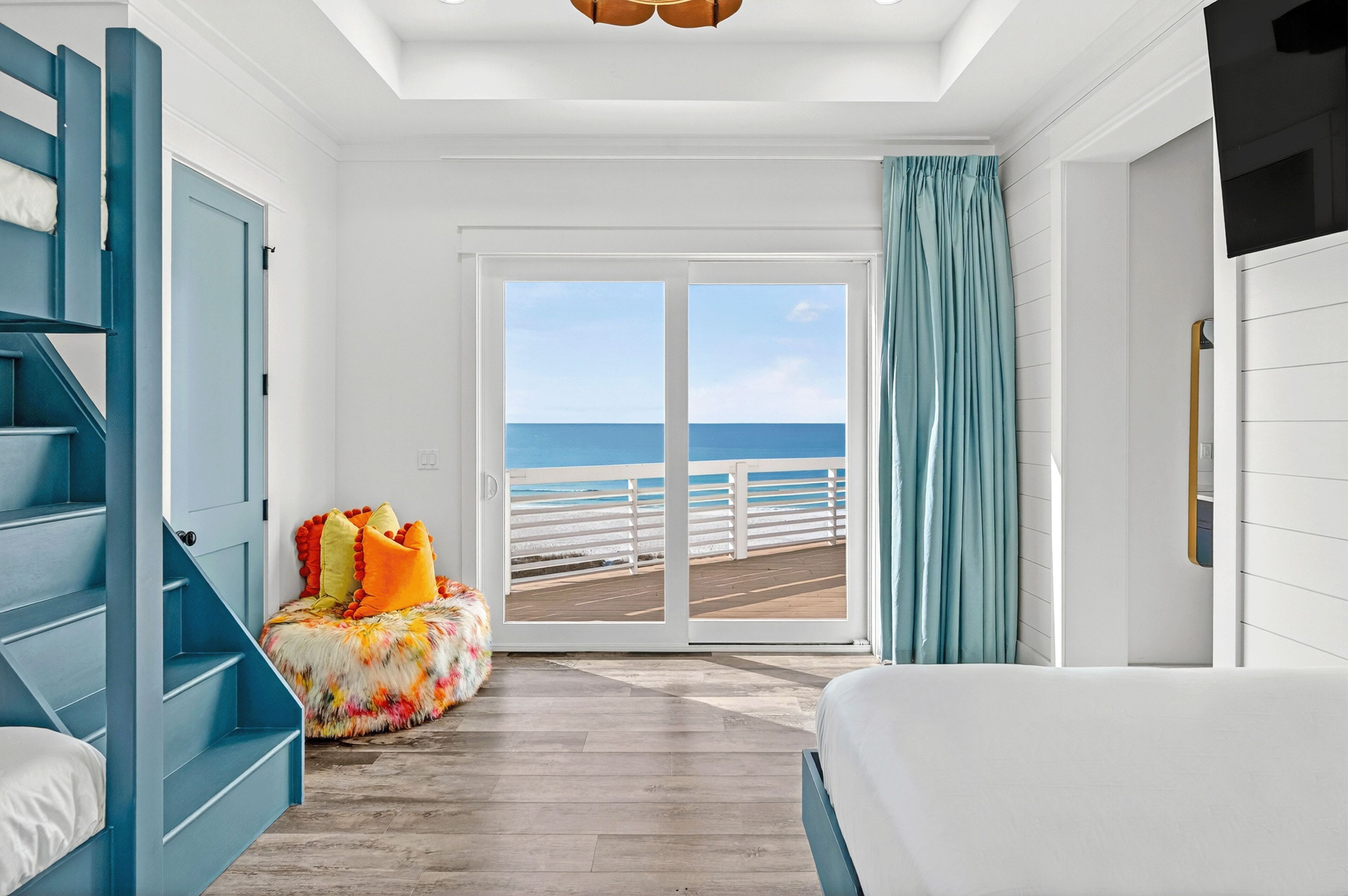A room with a view of the ocean, a blue staircase, a colorful ottoman, and a white bed.
