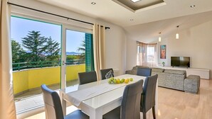 A modern living room with a dining area, a sofa, a TV, and a balcony with a view of trees and buildings. - Dionis apartment - 3 bedroom apartment with shared pool in a quiet neighborhood (Zadar)