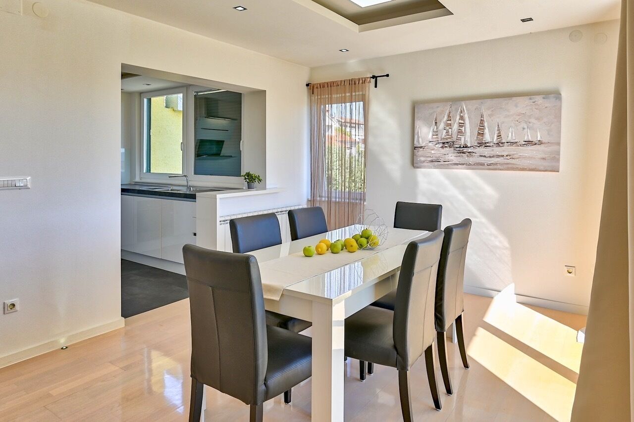 Un área de comedor moderna con una mesa blanca y sillas negras, una cocina al fondo y un cuadro de un velero en la pared.