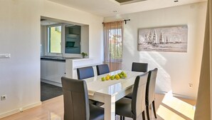 A modern dining area with a white table and black chairs, a kitchen in the background, and a sailboat painting on the wall. - Dionis apartment - 3 bedroom apartment with shared pool in a quiet neighborhood (Zadar)