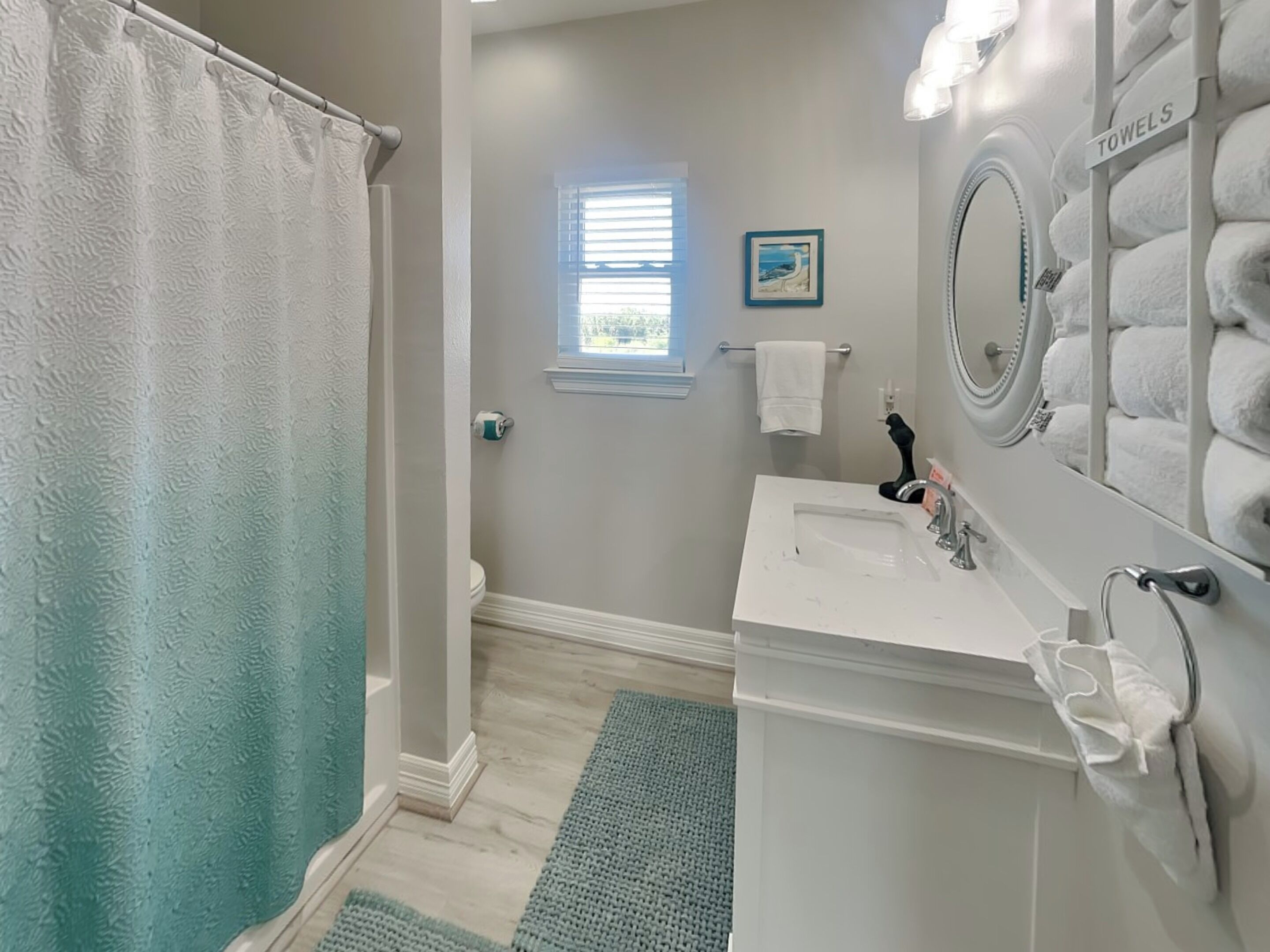 Une salle de bain équipée d’une douche, d’un lavabo, d’un porte-serviettes, d’une fenêtre avec des stores et d’un tableau encadré au mur.