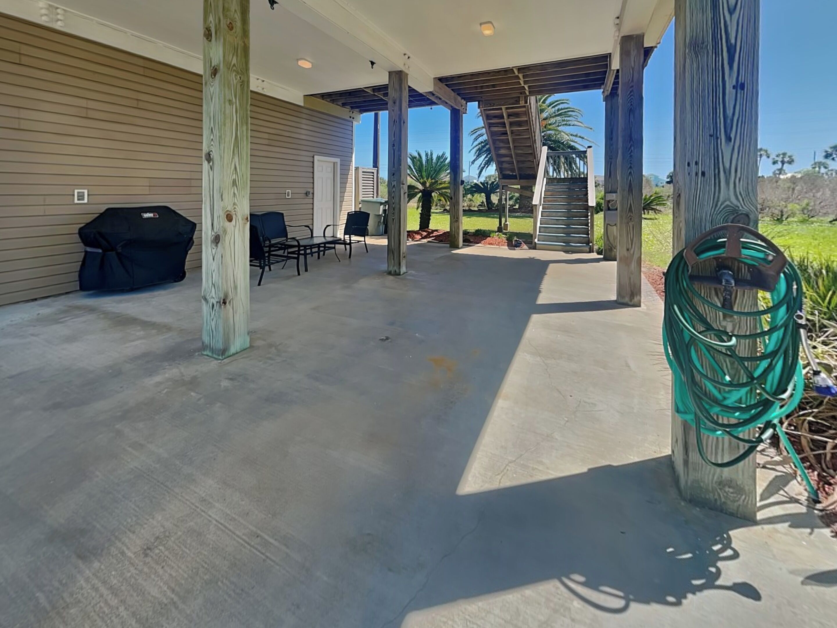 A covered patio area with a concrete floor, a barbecue grill, outdoor furniture, and a garden hose.
