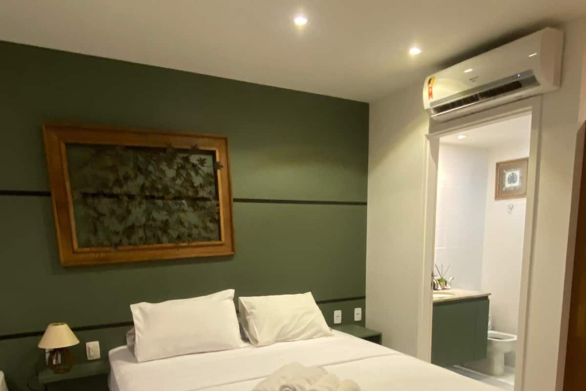 A hotel room with a bed, a nightstand, a painting, an air conditioning unit, and a bathroom visible through an open door.