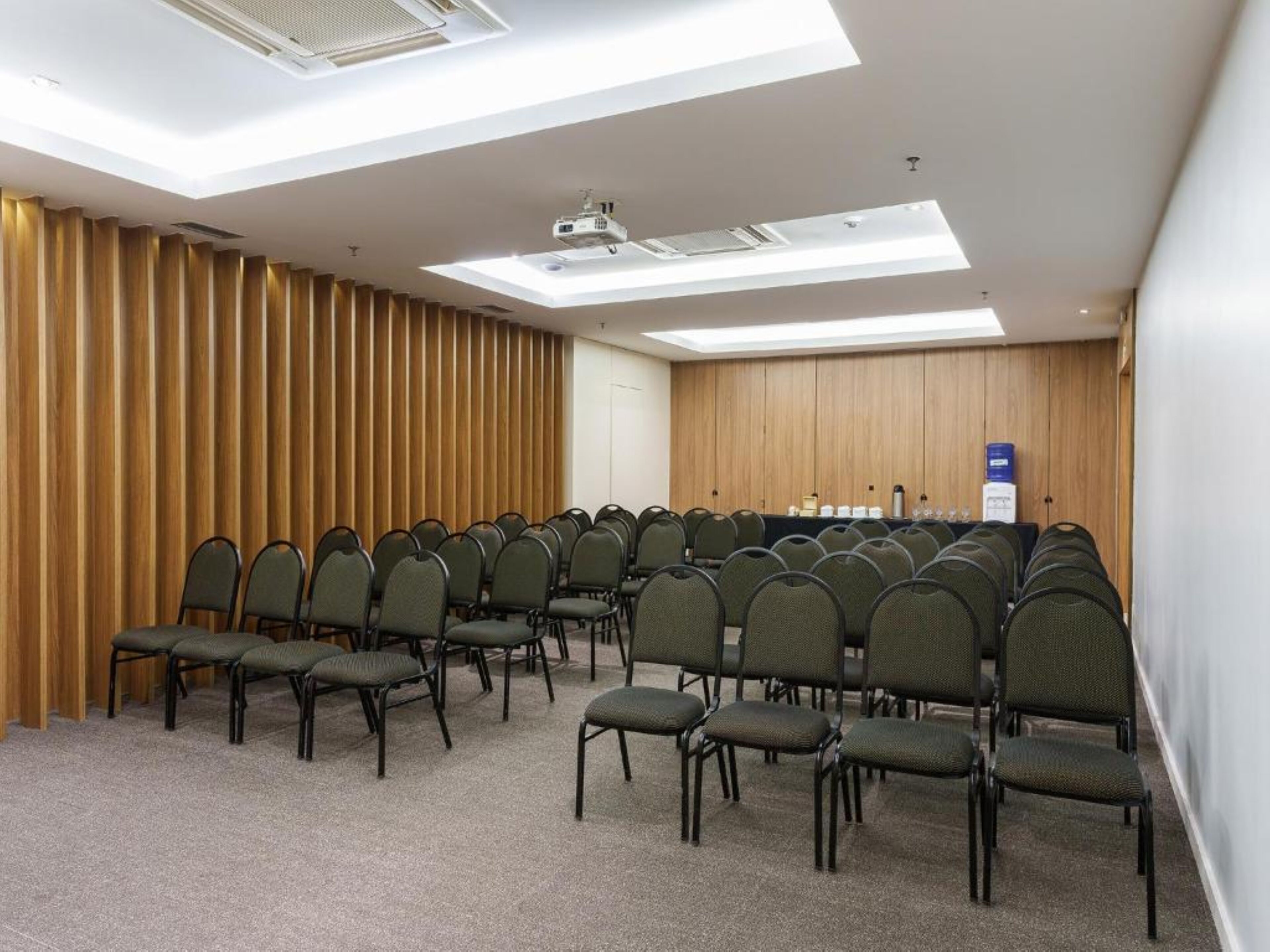 Une salle de conférence avec des rangées de chaises, un projecteur et un mur recouvert de panneaux en bois.