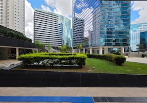 A modern urban plaza with tall buildings, a central landscaped area with bushes, and a paved walkway.