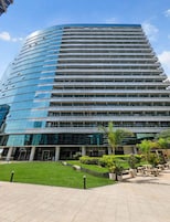 Un moderno edificio de gran altura con fachada de vidrio, un patio ajardinado con asientos y un cielo despejado.