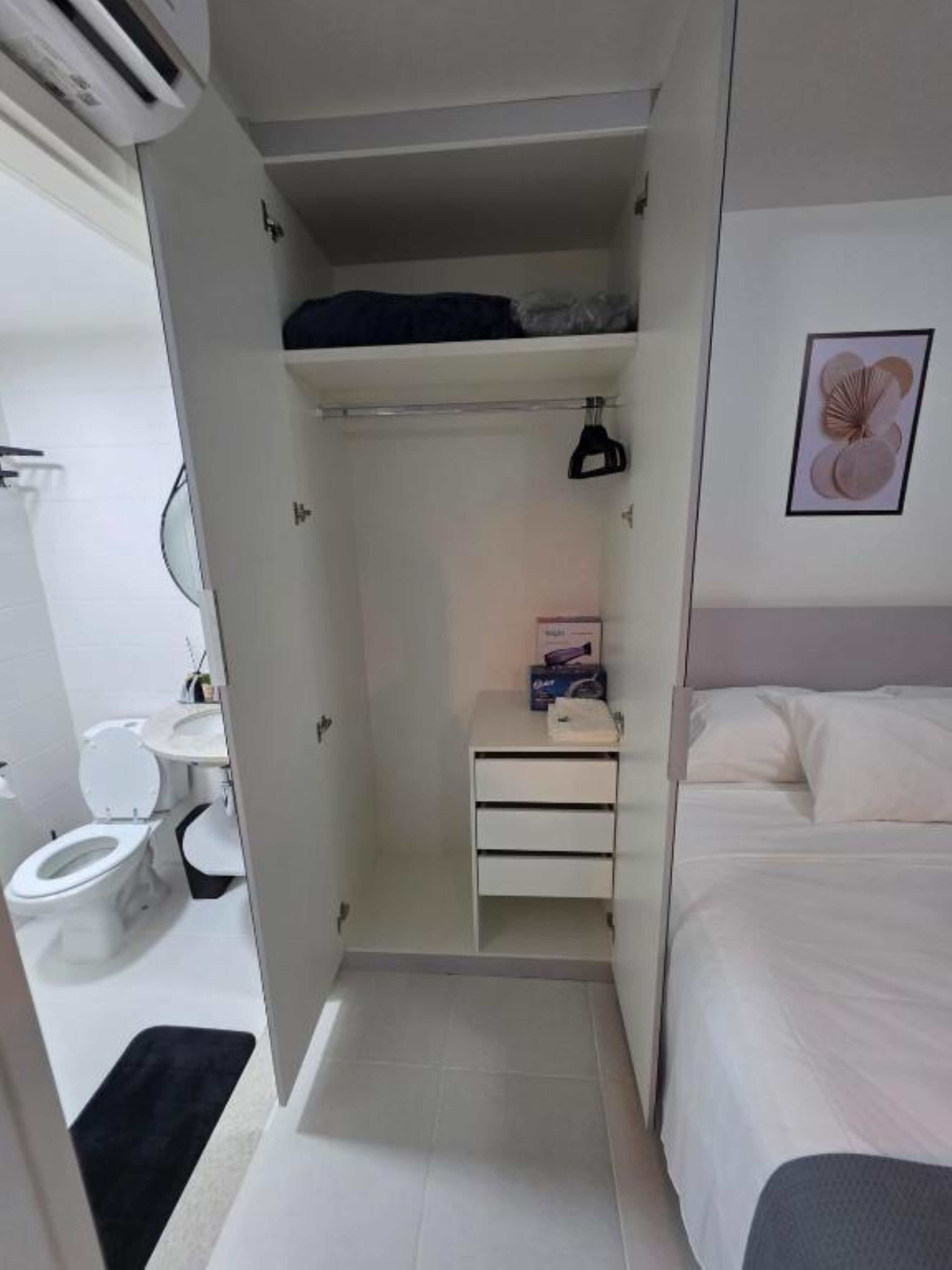 A compact hotel room with a bed, a small bedside table, a closet with hanging space, a toilet, and a sink.