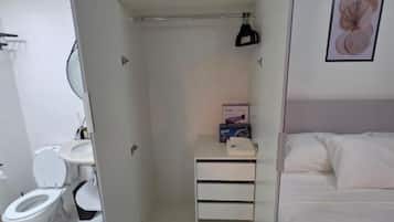 A compact hotel room with a bed, a small bedside table, a closet with hanging space, a toilet, and a sink.