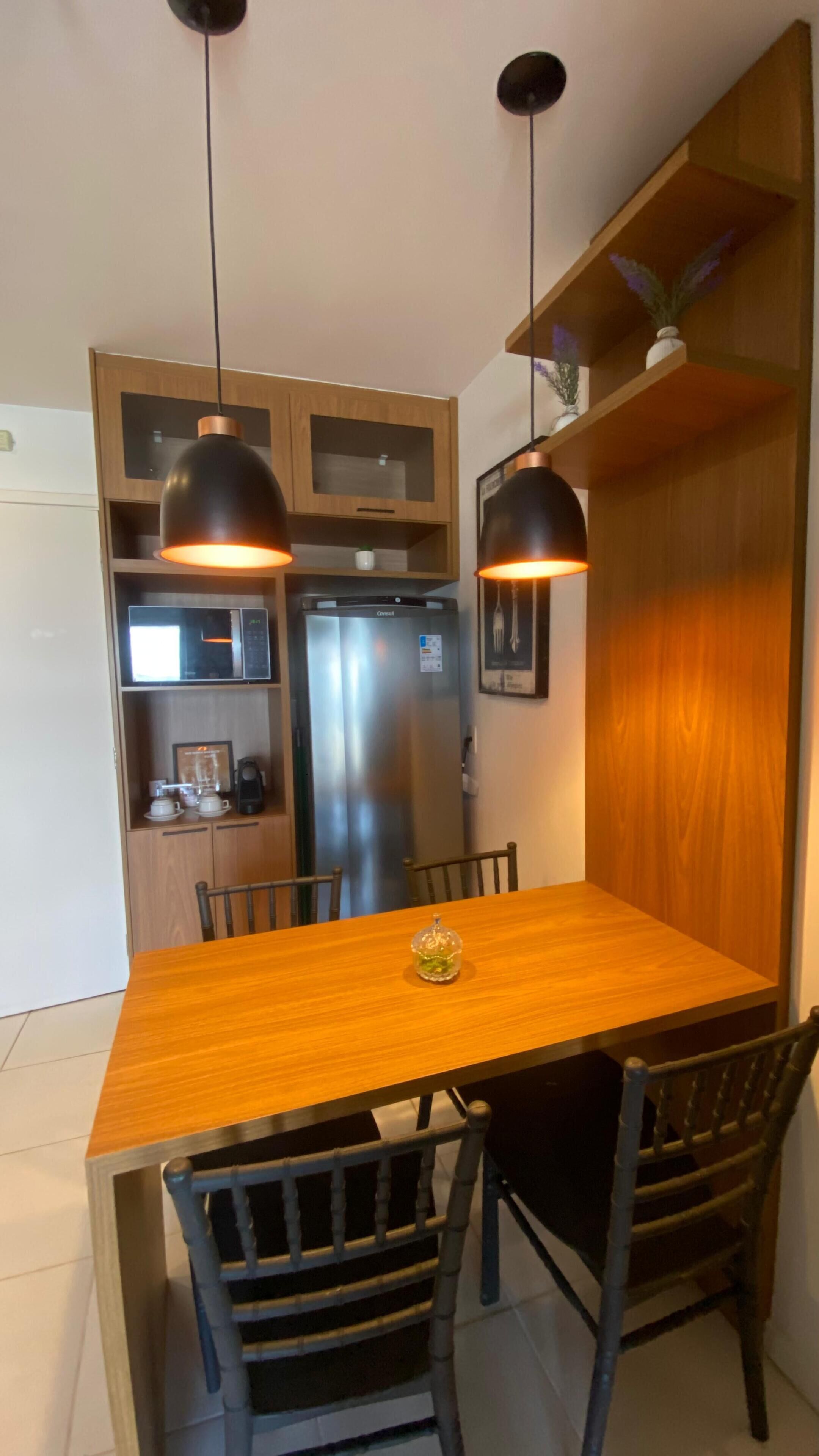 A compact kitchen with a wooden table, two chairs, a refrigerator, and pendant lights.