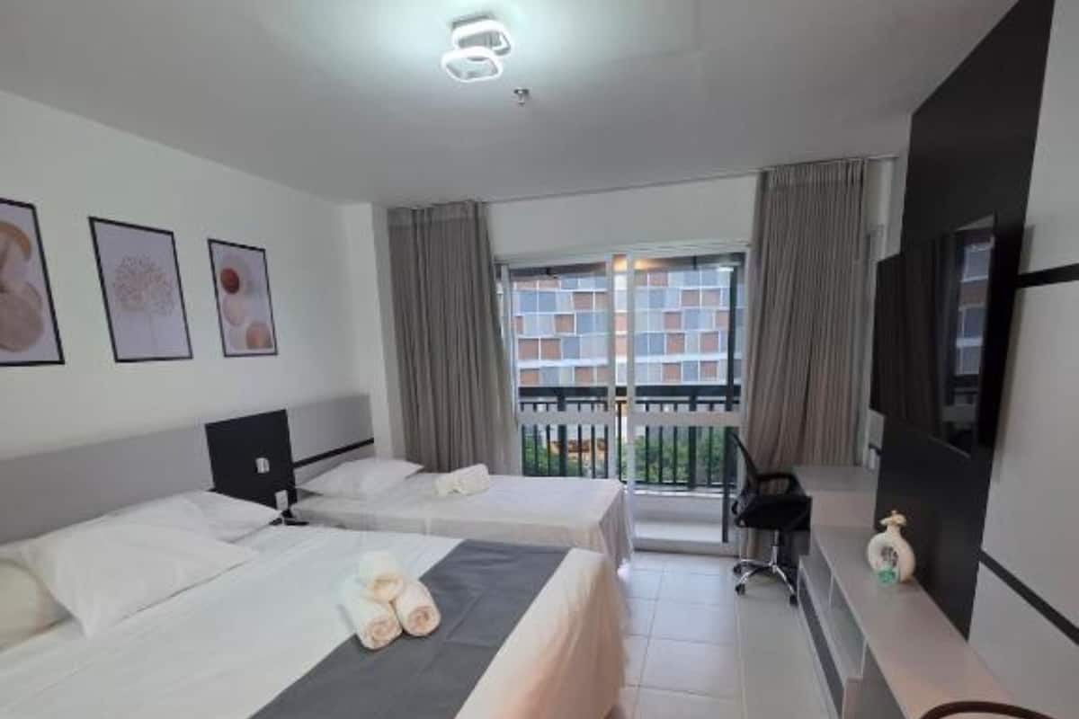 A hotel room with two beds, a desk, a chair, a TV, and a balcony with a view of buildings.