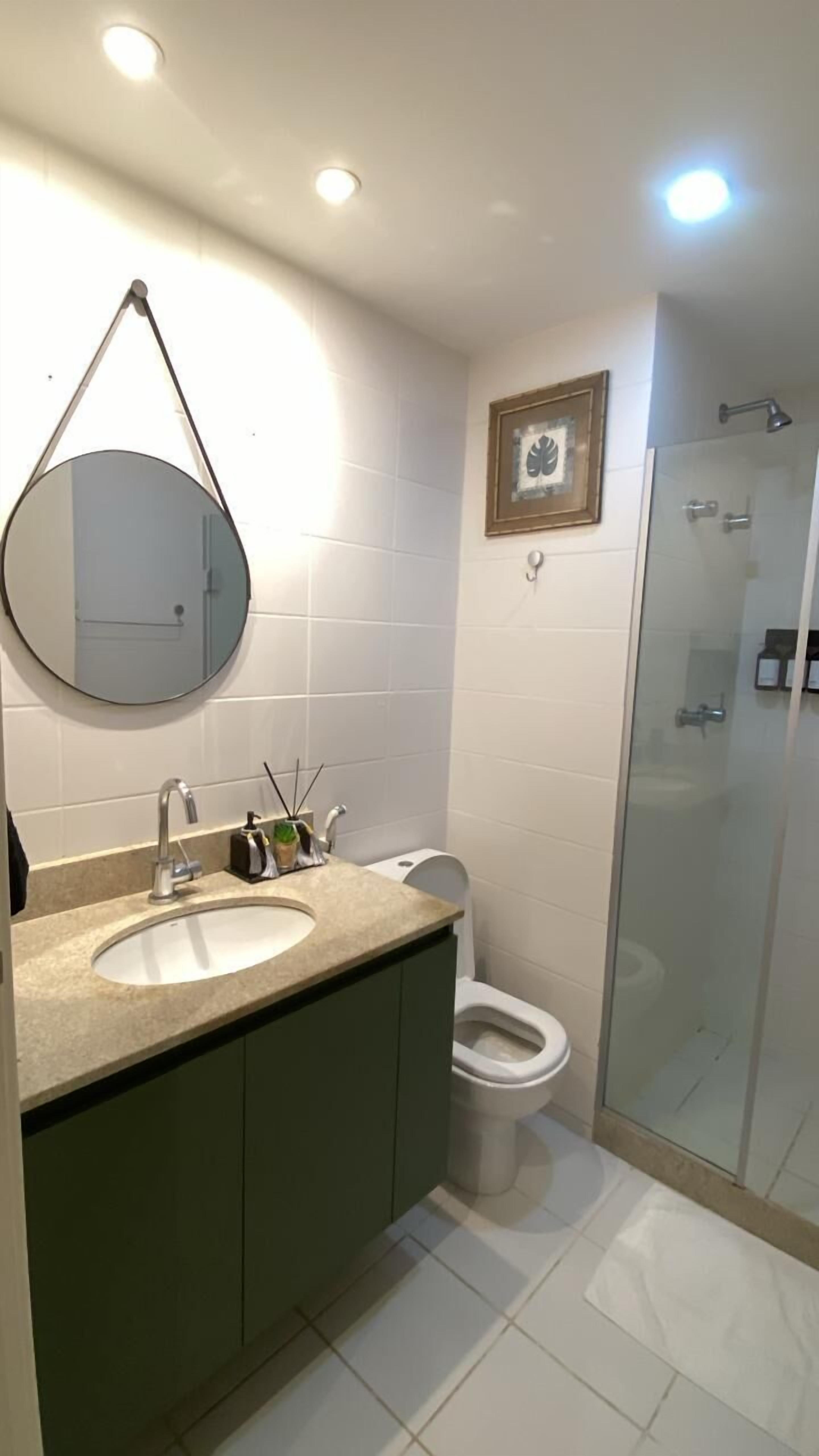 Kylpyhuoneessa on pyöreä peili, kaksi pesuallasta, wc ja lasiseinällä varustettu suihku.