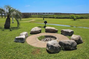 A circular stone fire pit surrounded by large rocks, with a palm tree and a wooden boardwalk in the background. - Pointe West 202 - The Hermit Hideaway (Galveston)