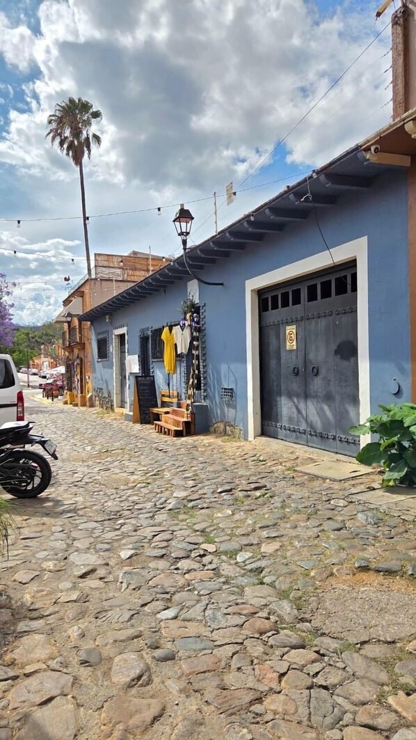 Eine Kopfsteinpflasterstraße mit einem blauen Gebäude, das eine große Garagentür, ein abgestelltes Motorrad am Rand und eine Palme im Hintergrund zeigt.
