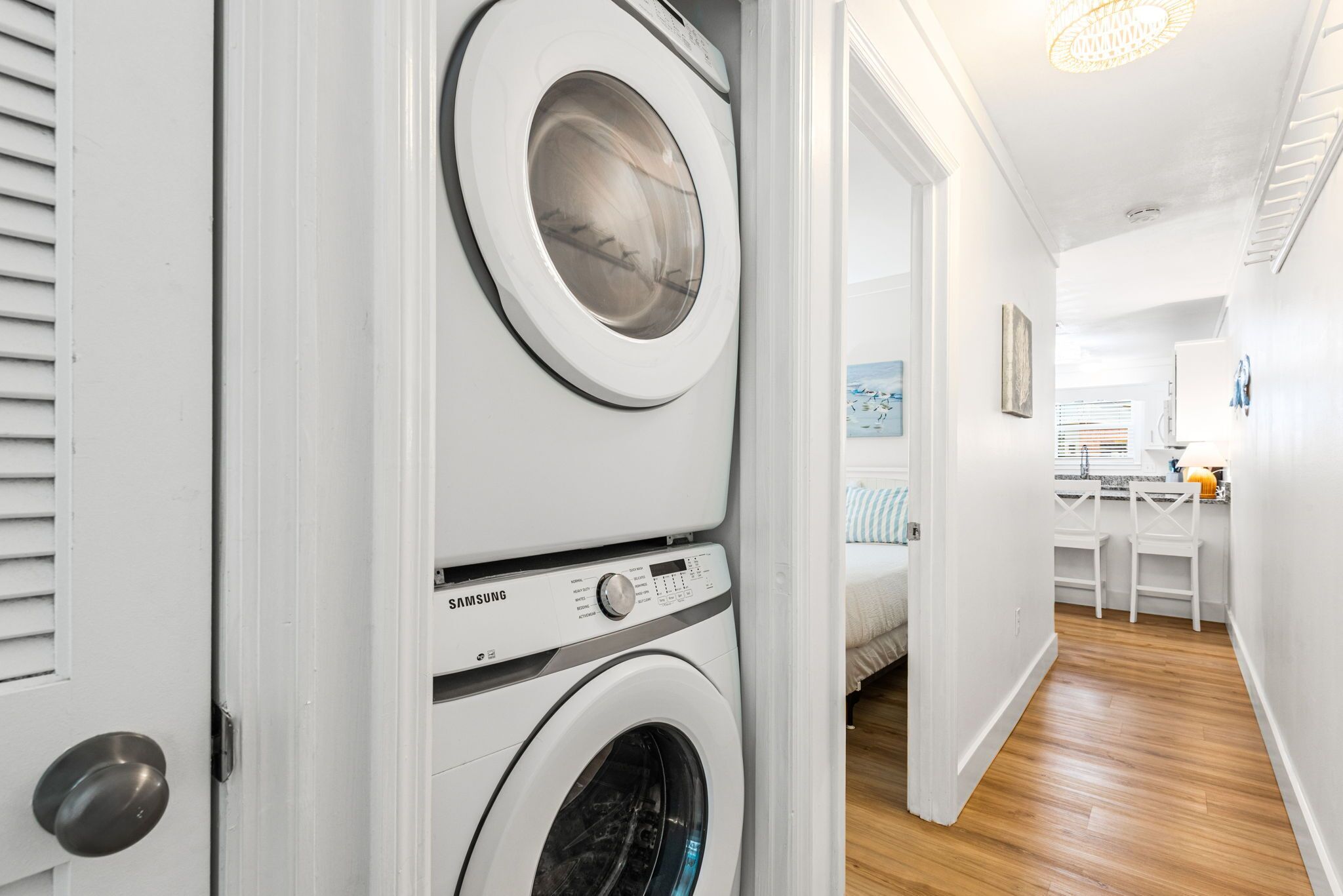 A laundry room with stacked washers and dryers, a wooden floor, and a hallway leading to a bedroom.