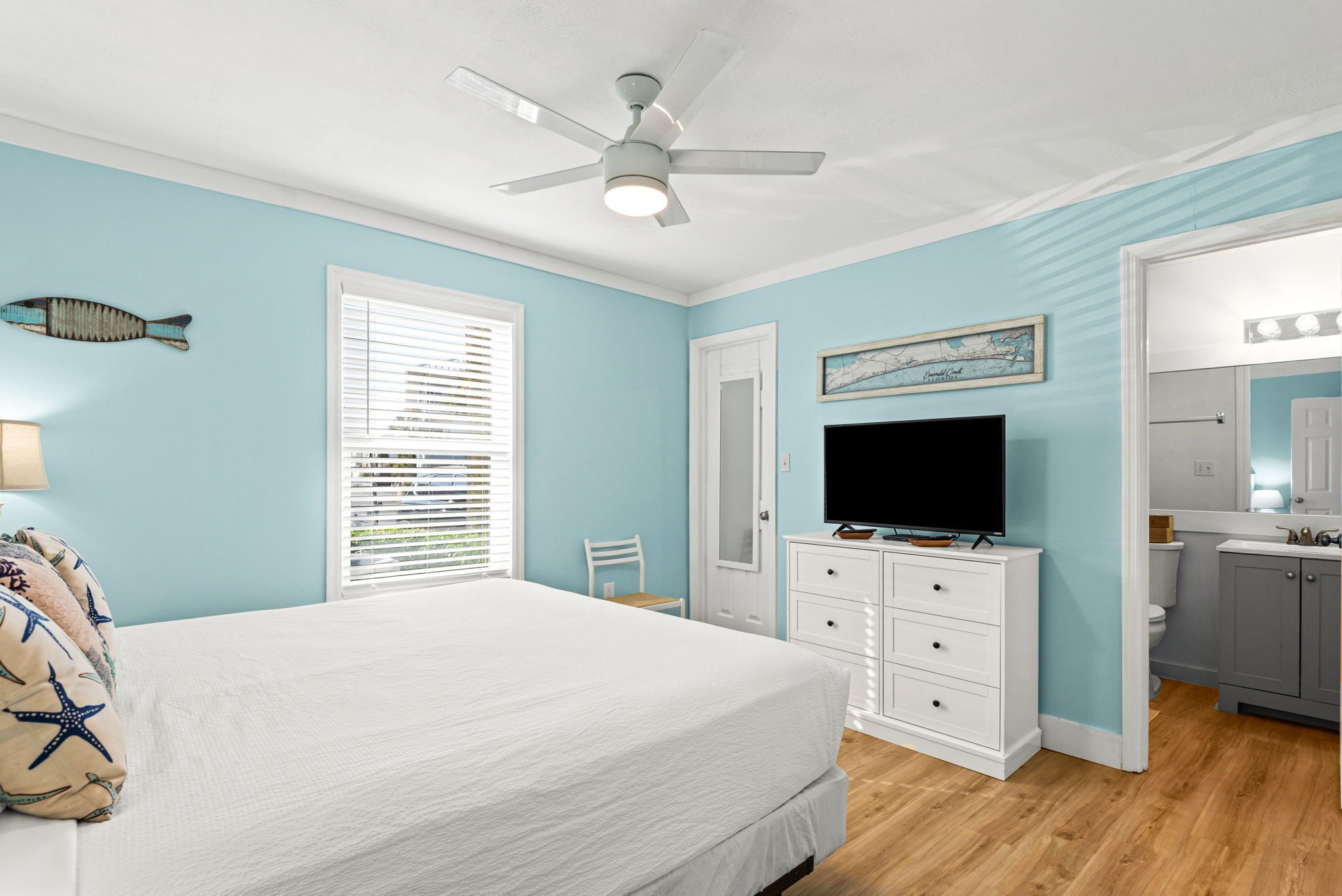A bedroom with a bed, a ceiling fan, a TV, a dresser, and a bathroom with a sink and mirror.