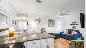 A modern kitchen with white cabinetry, a granite countertop, and a built-in stove. There is a dining area with a white table and chairs, and a living room with a blue sofa and a TV mounted on the wall. - Pet-friendly Corner 1st Floor Condo in Seagrove, Steps From the Beach (Santa Rosa Beach)