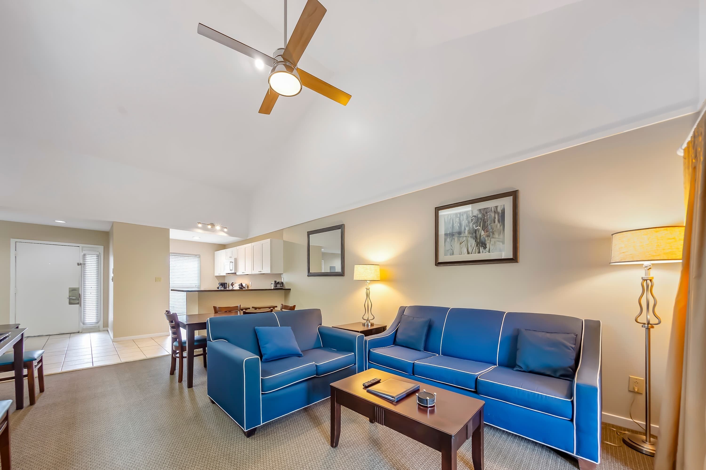 A living room with two blue sofas, a coffee table, a ceiling fan, a dining area, and a kitchen in the background.