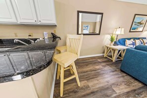 A kitchen with a black countertop and white cabinets, a wooden chair, a blue sofa, and a small table with a lamp. - New Bern Lakeside Getaway-Modern 2BR Apartment with Resort Amenities (New Bern)