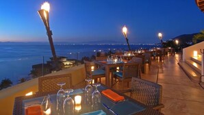An outdoor dining area with tables set for two, overlooking a body of water, with torches providing ambient lighting. - Garza Blanca Resort Junior Panoramic Suite in Puerto Vallarta 1 Bedroom, 1 Bath. (Puerto Vallarta)