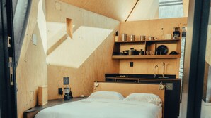 Ein kompakter, moderner Schlafraum mit Bett, Nachttischen, einer kleinen Sitzecke, Holzboden und einer Küchenzeile mit Regalen und Geräten.