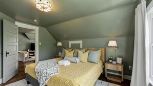 Ein Schlafzimmer mit schrägverdachter Decke, einem Bett mit gemustertem Überwurf, Nachttischlampen, einem Nachttisch und Blick auf einen Flur.