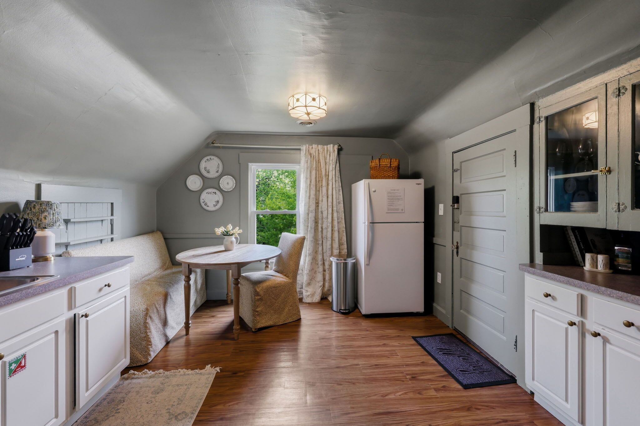 Een kleine keuken met witte kasten, een koelkast, een eettafel met stoelen en een raam met gordijnen.