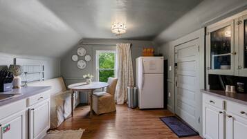 Een kleine keuken met witte kasten, een koelkast, een eettafel met stoelen en een raam met gordijnen.