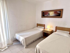 A hotel room with two single beds, a nightstand with a lamp, a painting on the wall, and a window with curtains. - Beachfront apartment with spectacular sea views (Segur de Calafell)