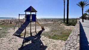 A playground with a slide and climbing structure, a sandy beach, and palm trees. - Beachfront apartment with spectacular sea views (Segur de Calafell)
