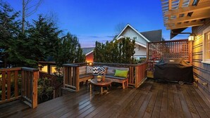 Eine Holzterrasse mit Bank und Tisch, umgeben von Bäumen und einem Gebäude.