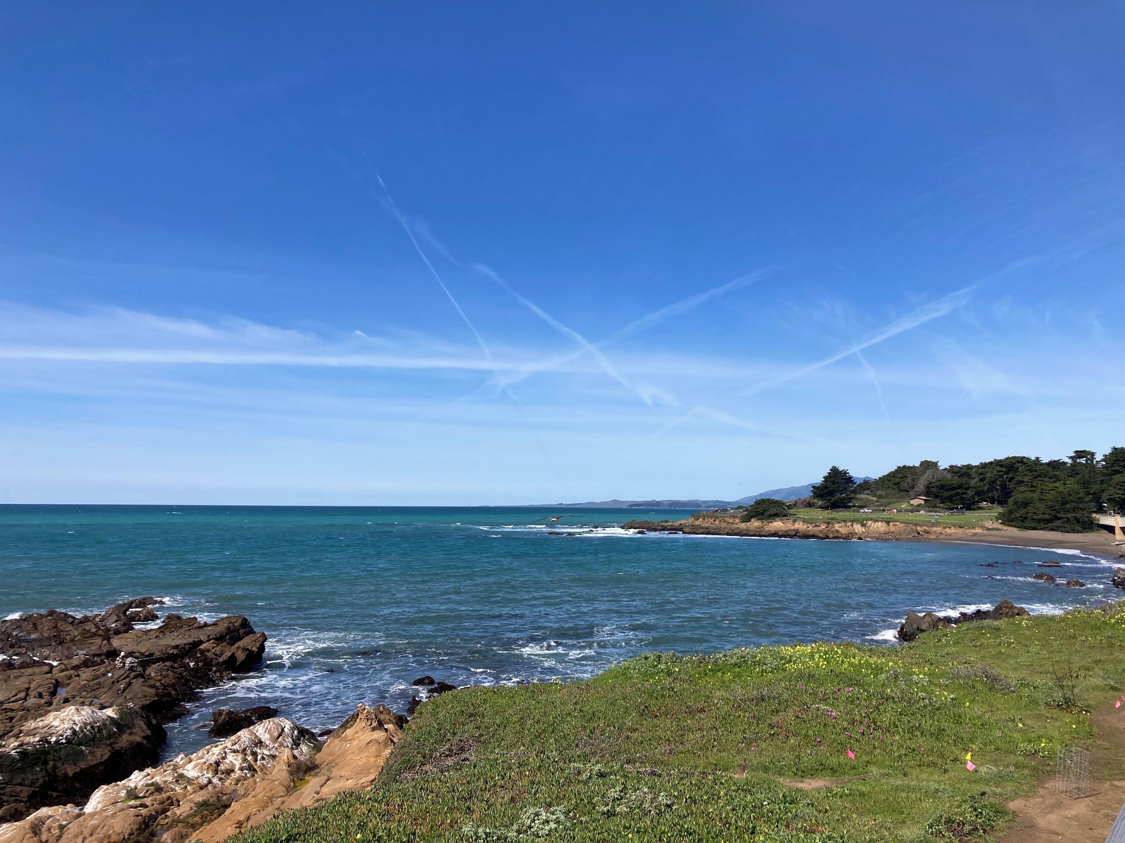 Un paysage côtier avec des rivages rocheux, un ciel bleu dégagé et des collines à l’horizon.
