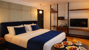 Quarto de hotel com cama, televisão, mesa de centro com café da manhã e estante de livros.