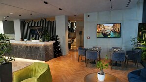 Um saguão de hotel com balcão de recepção, área de estar e uma televisão exibindo uma vista urbana.