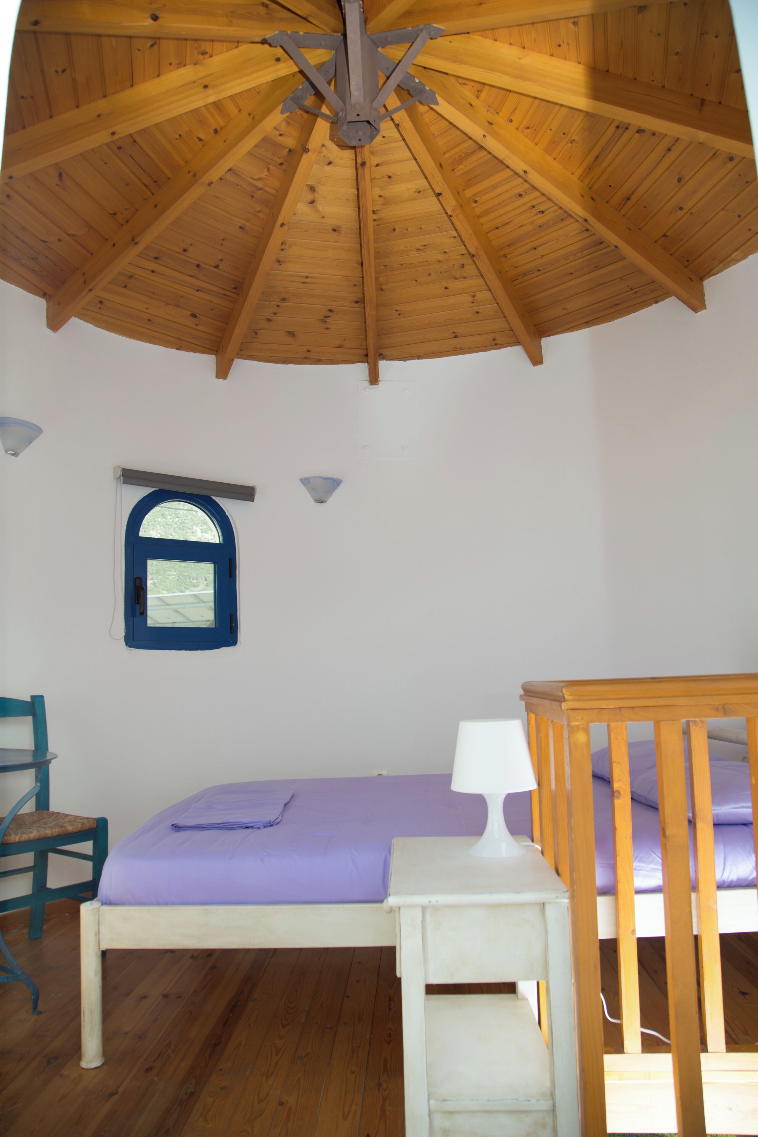 A room with a wooden ceiling, a bed with a purple bedspread, a white bedside table, a small window with a blue frame, and a wooden chair.