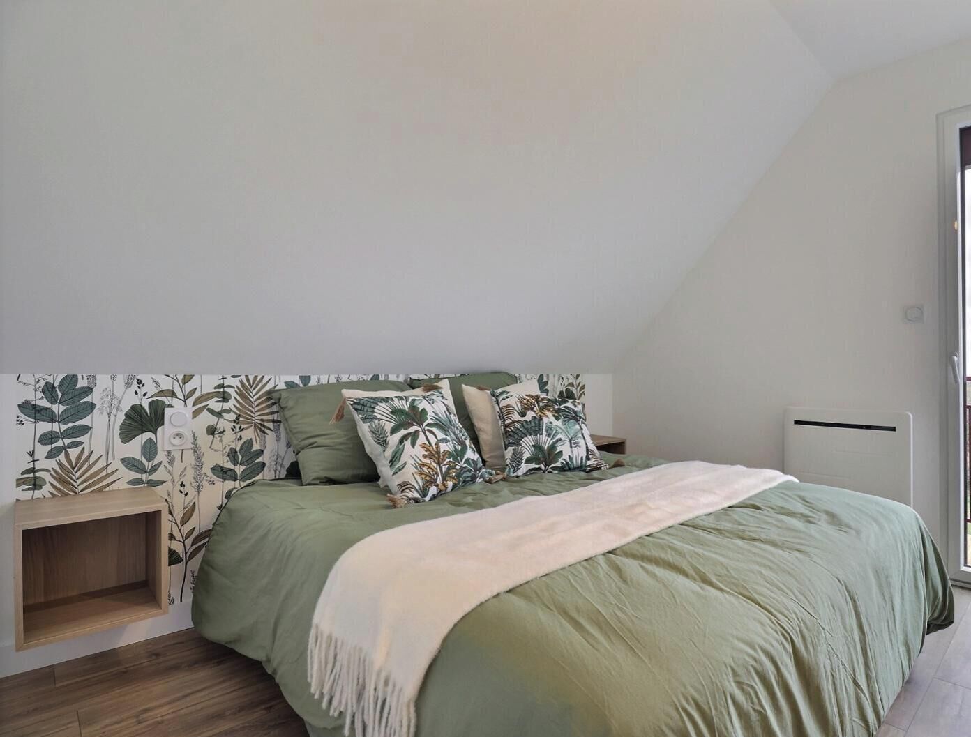 Un dormitorio con techo inclinado, cama con ropa de cama verde, manta blanca y pared con un estampado botánico.