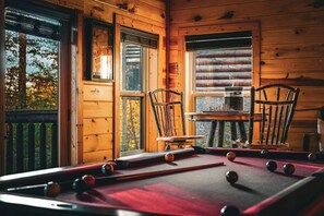 A pool table in a wooden cabin with two chairs, a window with blinds, and a view of trees. - NEW! Cozy 2BR Cabin w/PT, Hot Tub, & Pool Access (Sevierville)