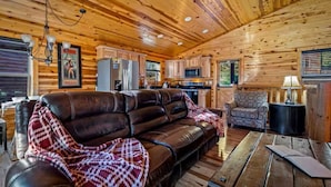 A log cabin interior with a leather sofa, patterned armchair, wooden dining table, and kitchen area. - NEW! Cozy 2BR Cabin w/PT, Hot Tub, & Pool Access (Sevierville)