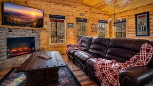 A log cabin interior with a fireplace, leather sofa, wooden walls, and a television. - NEW! Cozy 2BR Cabin w/PT, Hot Tub, & Pool Access (Sevierville)