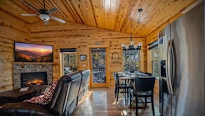A cabin interior with a fireplace, leather couch, dining area, and a large TV screen. - NEW! Cozy 2BR Cabin w/PT, Hot Tub, & Pool Access (Sevierville)