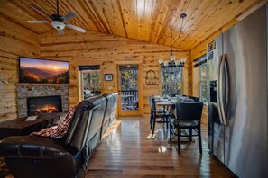 A cabin interior with a fireplace, leather couch, dining area, and a large TV screen. - NEW! Cozy 2BR Cabin w/PT, Hot Tub, & Pool Access (Sevierville)