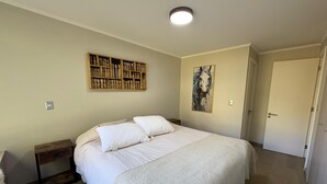 Un dormitorio con una cama, una mesita de noche, un estante en la pared, una puerta y un cuadro.