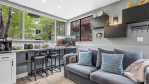 A modern kitchen with a dining area, a grey sofa, and a large window overlooking a balcony. - Cozy Mountain Nook-Tomboy Studio #133 (Telluride)