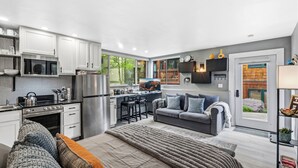 A modern kitchen with white cabinets, a stainless steel refrigerator, and a dining area with a gray sofa and a TV. - Cozy Mountain Nook-Tomboy Studio #133 (Telluride)