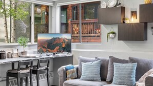 A modern living room with a large TV, a sofa with cushions, a desk with chairs, and a window with a view of mountains. - Cozy Mountain Nook-Tomboy Studio #133 (Telluride)